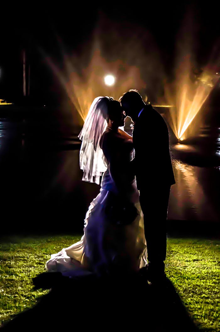 Contact Couple dancing romantically by the pond at night with dramatic lighting and water fountains.